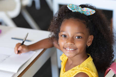 Portrait of smiling african american elementary schoolgirl writing on book at desk in classroom....