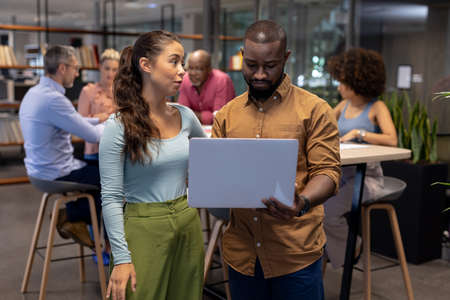 Young biracial businessman and businesswoman discussing over laptop against colleagues at workplace....