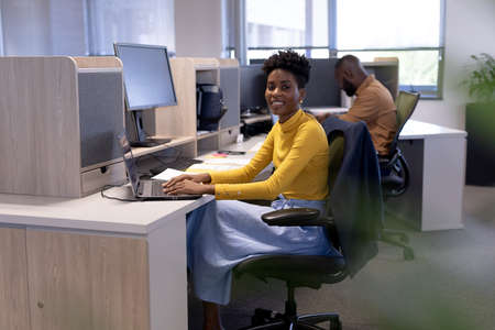 Portrait of smiling young african american female advisor using laptop at desk in modern workplace....