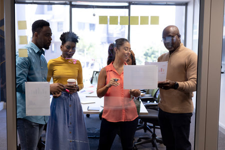 Multiracial male and female advisors planning strategy over documents seen through glass wall....
