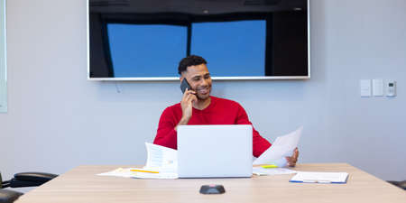 Smiling young hispanic businessman talking through smartphone while analyzing document in boardroom....