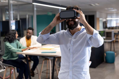 Smiling caucasian businessman wearing virtual reality simulator at modern workplace. unaltered,...