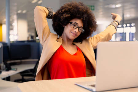 Tired young female african american advisor stretching while looking at laptop in workplace. unaltered, technology, business and modern office concept.の写真素材