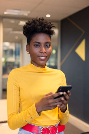 Portrait of smiling young african american female advisor holding smartphone at modern workplace....