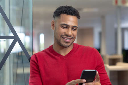 Smiling hispanic male advisor text messaging through smartphone leaning on glass wall in boardroom....