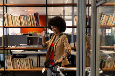 Happy young african american female advisor walking with bicycle while waving hand by bookshelf....