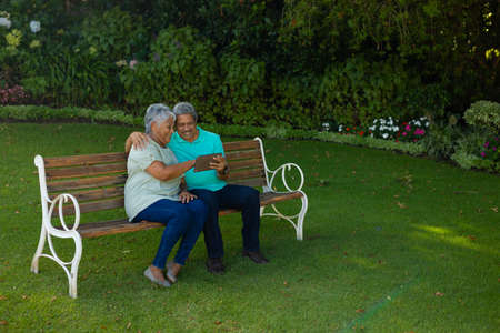 Smiling biracial senior couple using digital tablet while sitting on bench against plants in park. wireless technology, nature, unaltered, lifestyle, love, togetherness and retirement concept.の写真素材