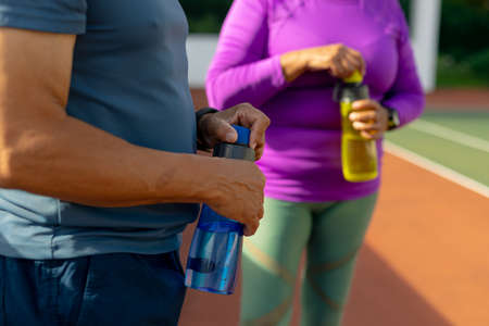 Midsection of biracial senior man and woman opening water bottles while standing in tennis court. unaltered, sport, thirsty, togetherness, love, retirement, healthy and active lifestyle concept.の写真素材