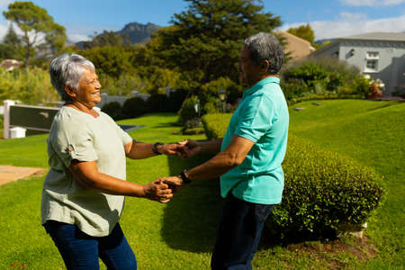 Side view of happy biracial senior couple holding hands and dancing in park during sunny day....
