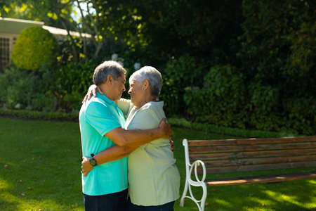 Happy biracial romantic senior couple looking at each other and embracing against plants in park....