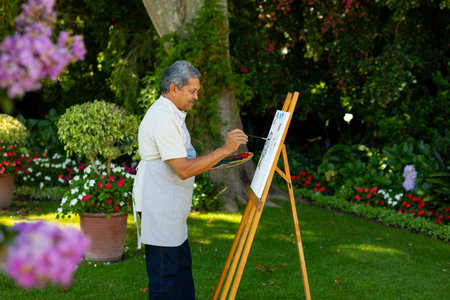 Side view of biracial senior man painting with watercolors on canvas against plants in yard. nature, palette, unaltered, lifestyle, painting, art, hobbies and retirement concept.の写真素材