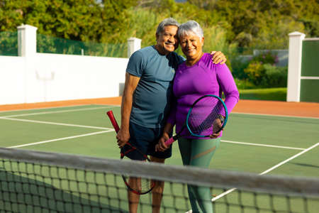 Portrait of happy biracial senior husband with wife holding rackets while standing in tennis court. unaltered, sport, competition, togetherness, love, retirement, healthy and active lifestyle.の写真素材