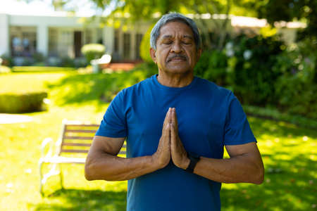 Biracial senior man with eyes closed meditating in prayer position against plants at yard. nature,...