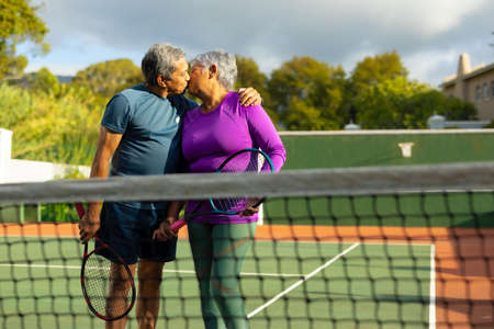 Biracial loving senior couple holding rackets kissing while standing in tennis court against sky. romance, unaltered, sport, togetherness, love, retirement, healthy and active lifestyle concept.の写真素材