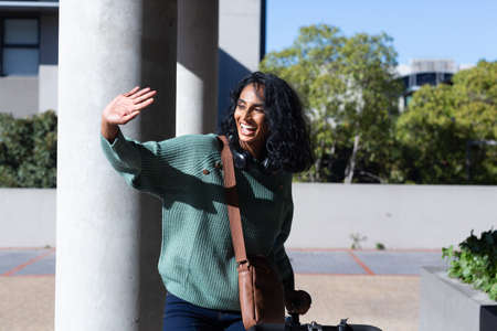 Cheerful young biracial businesswoman standing with bicycle while waving hand on sunny day....