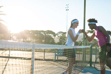 Happy multiracial young female tennis players giving fist bump over net at court. unaltered, sport, competition and tennis game concept.の写真素材