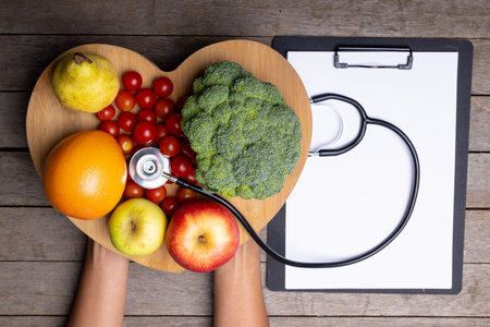 African american woman holding heart shape with vegetables, fruits, stethoscope by report on table. hand, clipboard, copy space, unaltered, healthy food, diet, medical, heart, disease and healthcare.の写真素材