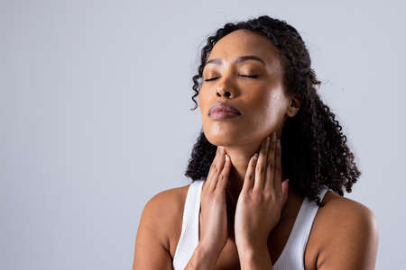 African american mid adult woman with eyes closed touching painful throat against white background. copy space, throat, thyroid, medical, pain, sickness and healthcare concept.の写真素材