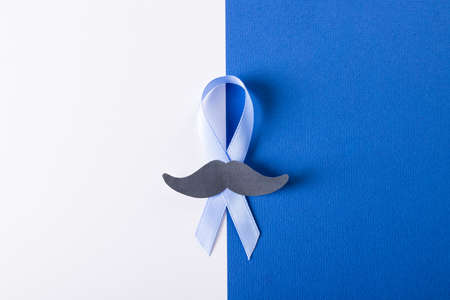 Overhead view of blue prostate cancer awareness ribbon with paper mustache on white, blue...