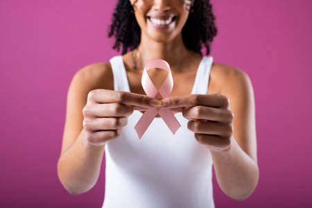 Midsection of smiling african american mid adult woman holding pink breast cancer awareness ribbon. pink background, breast, cancer, medical, ribbon, awareness, support, healthcare and alertness.の写真素材