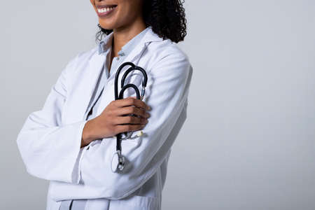 Midsection of smiling african american mid adult female doctor wearing lab coat holding stethoscope. white background, copy space, doctor, medical, healthcare, hospital and medical occupation.の写真素材