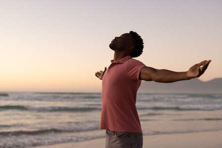 Side view of african american young man with arms outstretched standing against sea and clear sky. sunset, copy space, nature, unaltered, beach, lifestyle, enjoyment and holiday concept.の写真素材