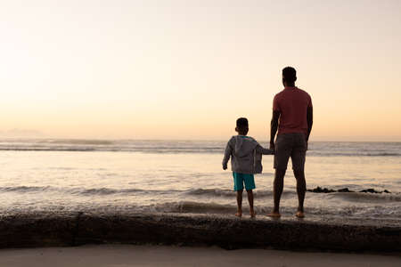 Rear view of african american young man with son looking at sea against clear sky during sunset. nature, unaltered, beach, childhood, family, togetherness, lifestyle, enjoyment and holiday concept.の写真素材