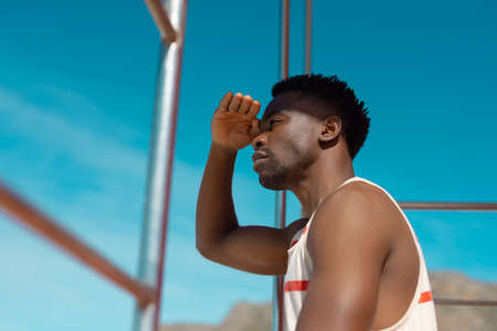 Side view of african american young man shielding eyes looking away while sitting against blue sky. copy space, summer, unaltered, beach, lifestyle, enjoyment and holiday concept.の写真素材