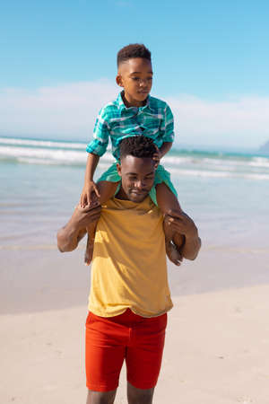 African american young father carrying son on shoulders while standing against sea and blue sky. copy space, nature, summer, unaltered, beach, childhood, family, togetherness, enjoyment, holiday.の写真素材