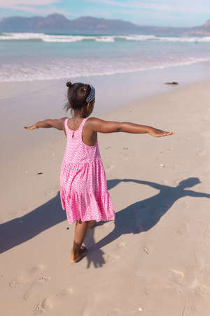 Rear view of playful african american girl with arms outstretched walking at beach against sky. summer, nature, childhood, unaltered, beach, lifestyle, enjoyment and holiday concept.の写真素材
