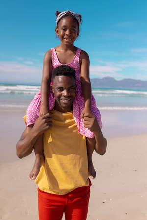 Portrait of smiling african american young man carrying daughter on shoulders against sea and sky....