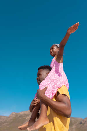 Low angle view of african american girl with arms outstretched sitting on young father&#39;s shoulders. sky, copy space, happy, unaltered, childhood, family, togetherness, enjoyment, holiday, beach.の写真素材