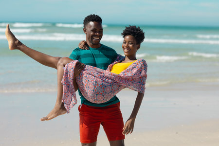 Happy african american young man carrying young girlfriend while standing at beach against sea. summer, nature, unaltered, beach, love, togetherness, lifestyle, enjoyment and holiday concept.の写真素材