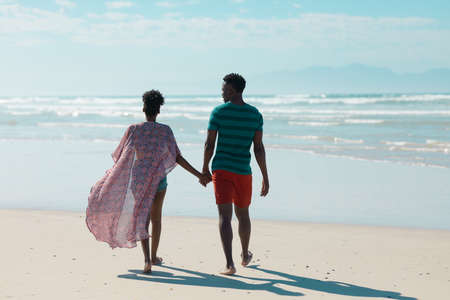 Rear view of african american young man holding girlfriend&#39;s hands and walking at beach against sky. nature, summer, unaltered, beach, love, togetherness, lifestyle, enjoyment and holiday concept.の写真素材