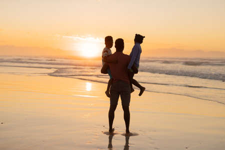 Rear view of african american young man picking up son and daughter on beach against sky at sunset. nature, unaltered, beach, childhood, family, togetherness, lifestyle, enjoyment and holiday.の写真素材