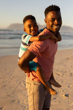 Portrait of smiling african american young man piggybacking son at beach against clear sky at...