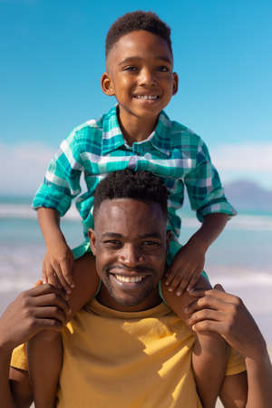Portrait of smiling african american young man carrying cute son on shoulders against blue sky. copy space, summer, unaltered, beach, childhood, family, togetherness, lifestyle, enjoyment, holiday.の写真素材