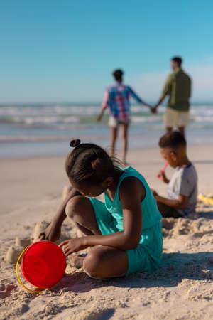 African american girl with pail making sand castle at beach against sky with family in background....