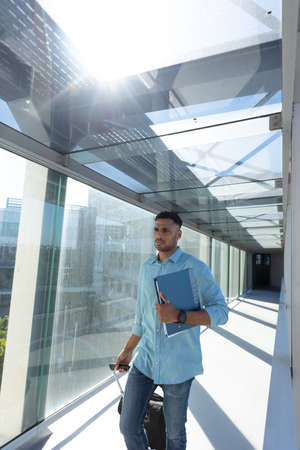 Young african american businessman walking with file and luggage at airport corridor on sunny day....