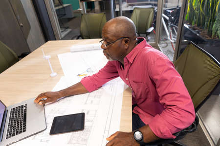 Mature african american engineer using laptop while sitting with blueprint at desk in workplace....