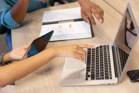 Cropped image of biracial colleagues discussing over digital tablet and laptop at desk in workplace....