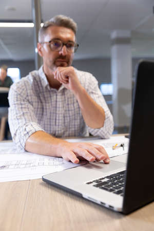 Caucasian male engineer using laptop while sitting with blueprint at desk in modern workplace. unaltered, creative business, technology and modern office concept.の写真素材
