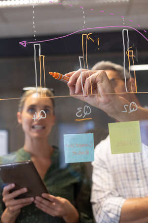 Caucasian businessman drawing bar graph on glass wall while discussing with businesswoman....