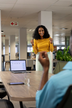 Happy biracial businesswoman cheering while looking at african american businessman at workplace. unaltered, business, technology, teamwork and modern office concept.の写真素材