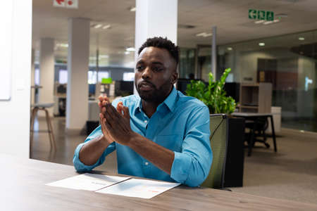 Confident african american businessman sitting with hands clasped and document at desk in workplace....