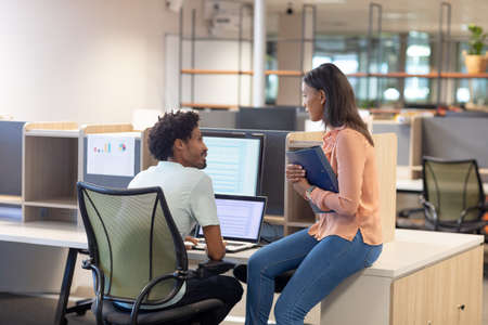 Biracial business colleagues discussing together while sitting at modern workplace cubicle....