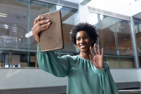 Smiling african american businesswoman waving on video call through tablet pc at workplace....