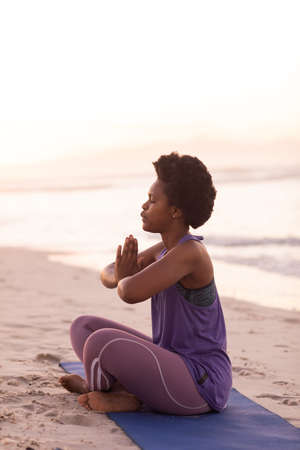 Side view of african american mature woman with afro hair meditating while sitting on mat at beach....