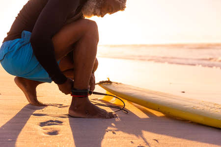 Bearded african american senior man tying surfboard string at beach against clear sky at sunset. copy space, nature, unaltered, retirement, aquatic sport, holiday and active lifestyle concept.の写真素材