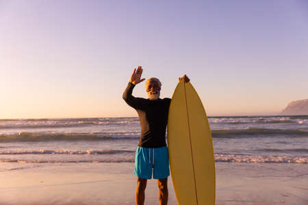 Bearded african american senior man with surfboard waving while standing against sea and clear sky....